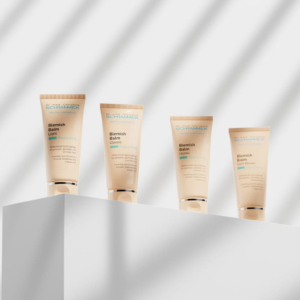 Vier tubes Dr. med. Christine Schrammek Blemish Balm in verschillende tinten op een witte display tegen een licht geblokte achtergrond.