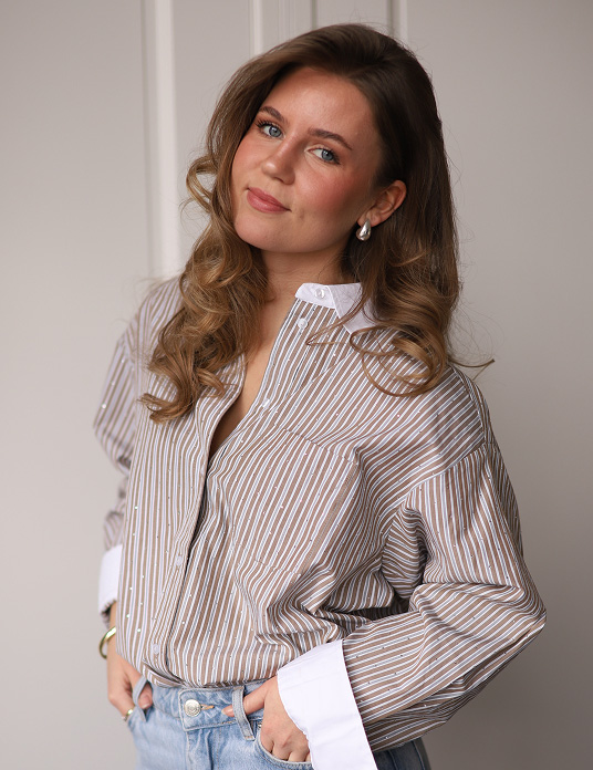 Celine de Oude met lang golvend haar draagt een gestreept overhemd en spijkerbroek terwijl ze ontspannen poseert.